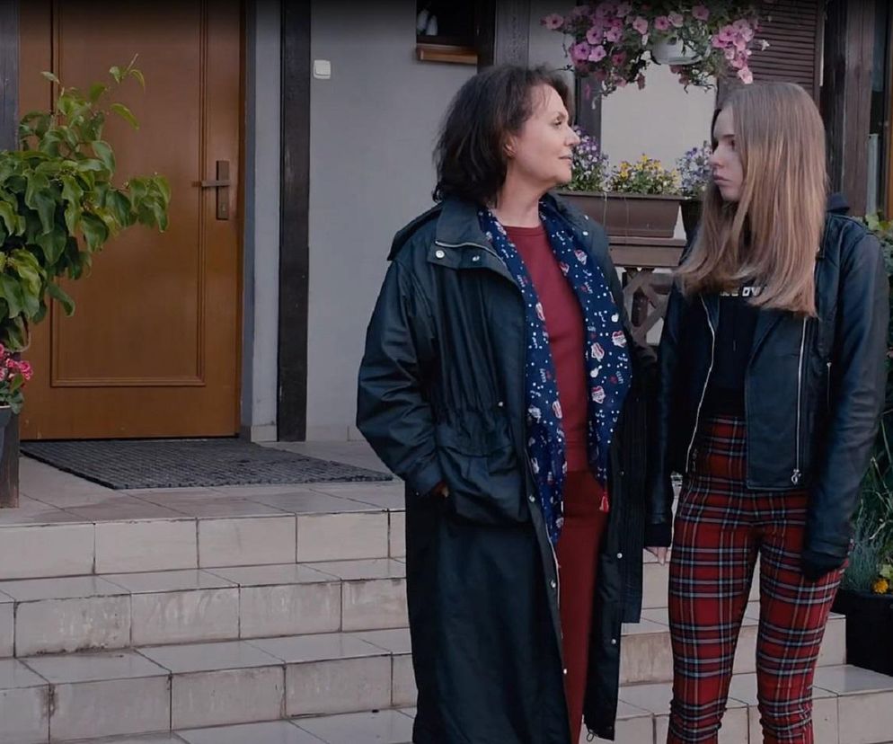 Małgorzata Pieńkowska jako Marysia Rogowska w długiej czarnej kurtce z niebieskim szalem i Nadia Smyczek jako Sara w czarnej ramonesce i spodniach w czerwoną kratę stoją przed domem. Marysia patrzy na Sarę, która spuszcza wzrok. Scena z serialu M jak miłość. Więcej informacji o serialu znajdziesz na Super Seriale.