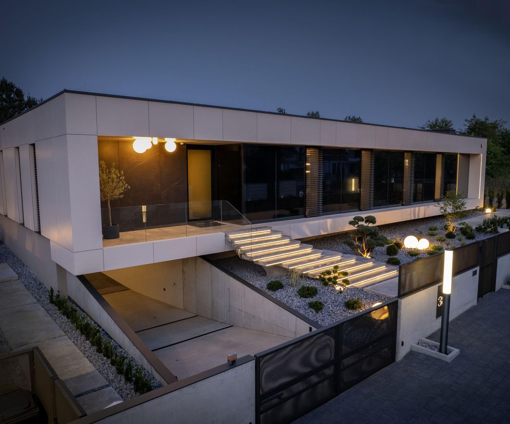 Nowoczesny dom jednorodzinny The Frame autorstwa Daniela Cieślika z podziemnym garażem, widoczny wieczorem z podświetlanymi schodami prowadzącymi do wejścia. Budynek posiada elewację z czarnego szkła i jasne ściany, a otoczenie zdobią ozdobne rośliny. Więcej o takich realizacjach przeczytasz na Architektura Murator Plus.