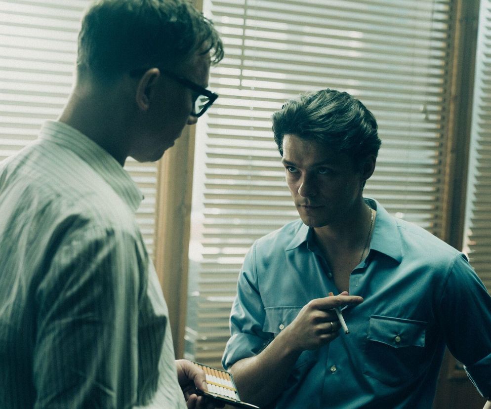 Tomasz Ziętek jako Igor Bielik w serialu „Czarna śmierć” na tle okna z żaluzjami, w intensywnej rozmowie z redaktorem naczelnym. Aktor w turkusowej koszuli, z papierosem w dłoni, patrzy z determinacją. Więcej o produkcji na portalu Super Seriale.