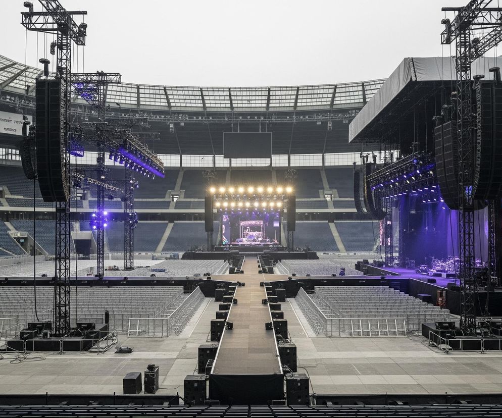 Wnętrze dużego, pustego stadionu sportowego widziane z trybun, z centralnie umiejscowioną sceną koncertową na drugim końcu boiska. Pośrodku sceny świecą intensywnie żółte reflektory, a po jej bokach widoczne są duże ekrany LED wyświetlające obrazy. Do sceny prowadzi długi, wąski podest otoczony rzędami pustych krzeseł i barierek ochronnych. Z boku sceny oraz wzdłuż podestu rozmieszczone są wysokie, metalowe konstrukcje z licznymi czarnymi głośnikami oraz niebiesko świecącymi reflektorami, rozświetlającymi ciemną przestrzeń pod zadaszeniem stadionu.