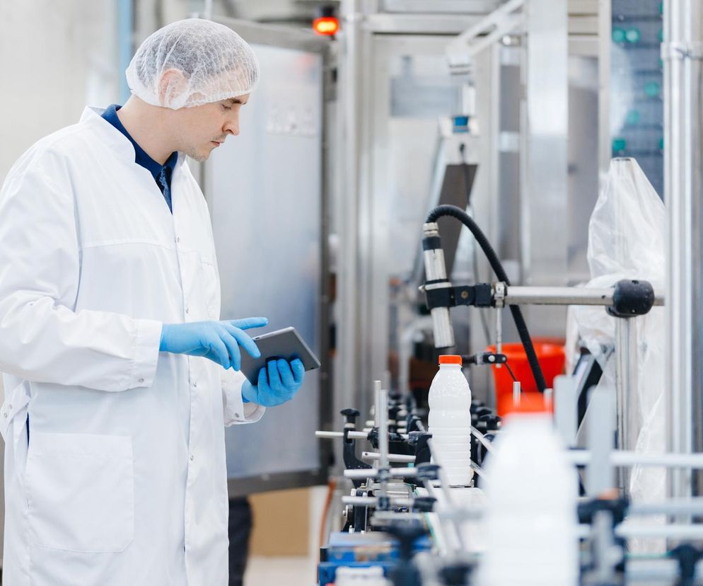 Pracownik mleczarni w białym fartuchu i czepku na linii produkcyjnej butelek mleka, kontroluje proces na tablecie. Symbolizuje upadek zakładu mleczarskiego w Lidzbarku Welskim, o czym szerzej można przeczytać na Super Biznes.