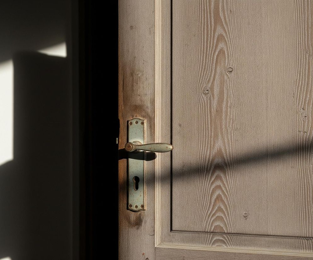 Uchylone drewniane drzwi z widocznym usłojeniem zajmują prawą część kadru. Na drzwiach zamontowana jest zielonkawa klamka z odrapanym metalowym szyldem i otworem na klucz. Po lewej stronie widać fragment ściany w jasnym odcieniu, na którą padają silne cienie, tworząc geometryczne kształty i ciemne plamy. Całość oświetlona jest ciepłym, bocznym światłem, tworzącym kontrastowe obszary i podkreślającym fakturę drewna.