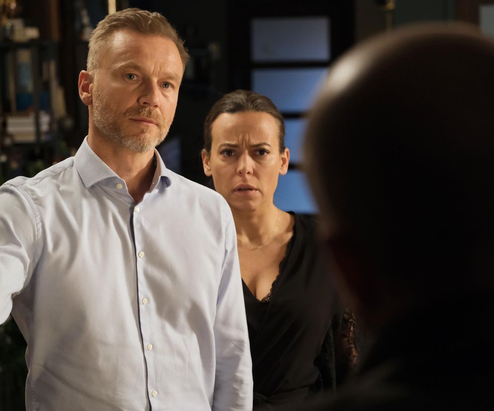 Andrzej Budzyński (Krystian Wieczorek) zaniepokojony patrzy na rozmówcę, podczas gdy w tle stoi przestraszona Magda Budzyńska (Anna Mucha). Oboje są w szoku, co widać na ich twarzach. Więcej o ich losach na Super Seriale.