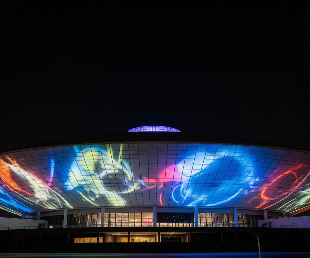 W centrum kadru wznosi się duży, eliptyczny budynek o futurystycznym kształcie, oświetlony od zewnątrz projekcjami świetlnymi. Fasada obiektu pokryta jest szklanymi panelami tworzącymi siatkę, na których wyświetlane są dynamiczne, abstrakcyjne wzory w jaskrawych kolorach: niebieskim, czerwonym, żółtym i fioletowym. Nad główną bryłą, na szczycie dachu, widoczna jest niewielka kopuła świecąca fioletowym światłem. Dolna część budynku, poniżej oświetlonej fasady, ma ciemne, przeszklone powierzchnie, za którymi widać słabe światło wnętrza. Całość otacza nocne, ciemne tło z kilkoma drobnymi, niewyraźnymi światłami w oddali po lewej stronie.