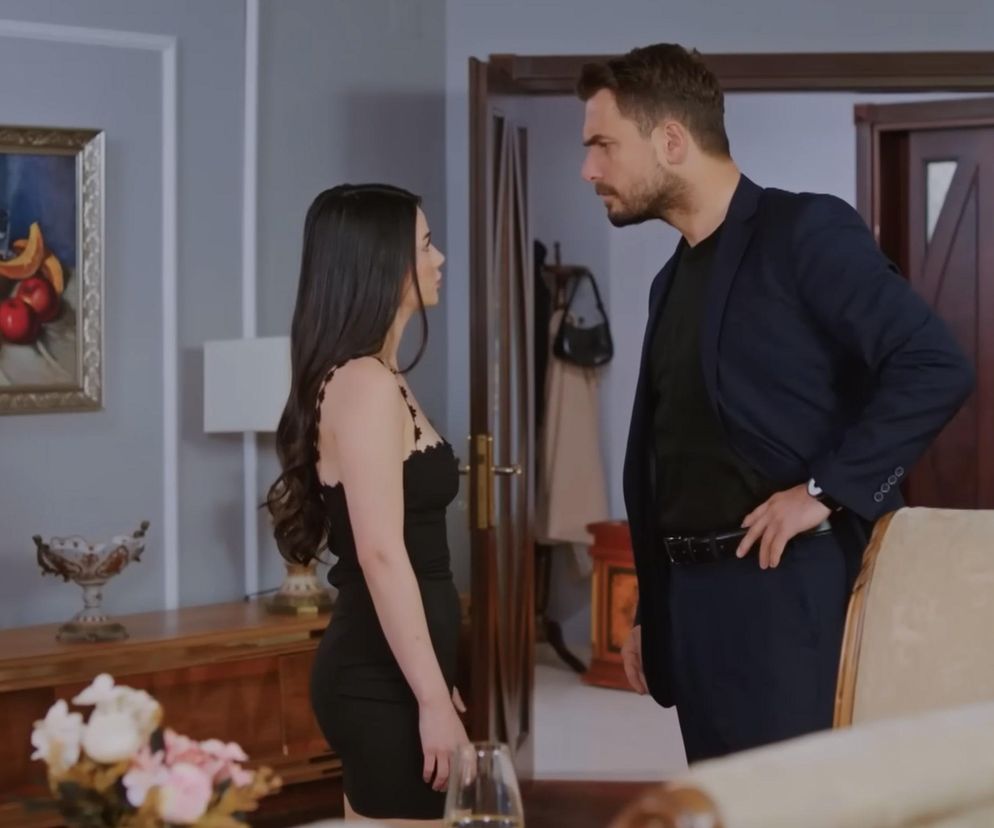 Bahar w czarnej sukience spogląda na Kuzeya, ubranego w granatową marynarkę, stojących w eleganckim salonie. W tle widać obraz z owocami oraz lampę. Scena przedstawia napięcie między bohaterami serialu „Miłość i nadzieja”. Więcej szczegółów na temat ich losów znajdziesz na Super Seriale.