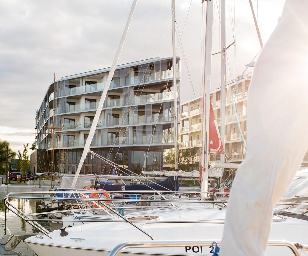 Nowoczesny budynek CLUBHOUSE w Szczecinie, z przeszkloną fasadą i balkonami, sfotografowany z perspektywy mariny, gdzie zacumowane są białe jachty, z promieniem słońca wpadającym w kadr. Więcej o architekturze nad wodą przeczytasz na Architektura Murator Plus.