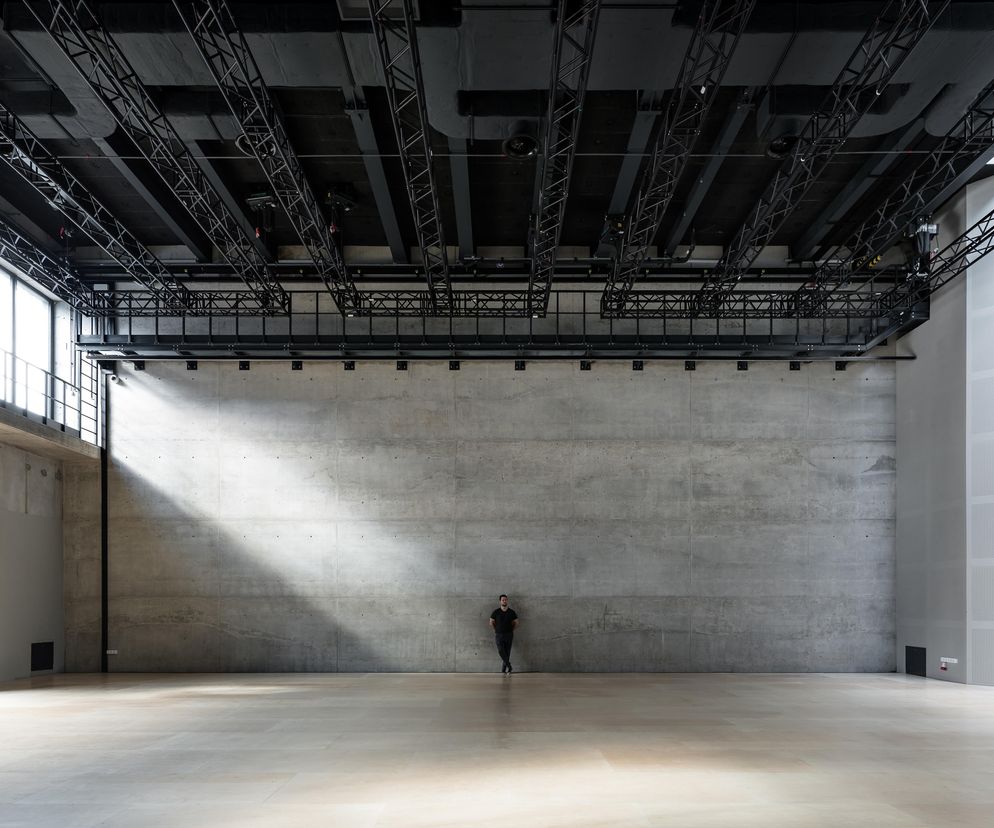 Pusta sala studyjna Polskiego Teatru Tańca w Poznaniu z człowiekiem opierającym się o betonową ścianę. Widoczne industrialne elementy: odkryta konstrukcja stalowa na suficie, surowy beton ścian. Na podłodze drewniane panele. O tym projekcie przeczytasz więcej na portalu Architektura Murator Plus.