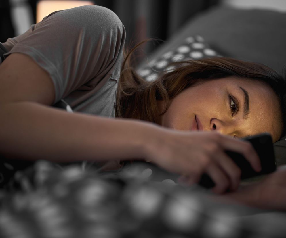 Kobieta leżąca w łóżku wieczorem, patrząca na smartfon, którego ekran oświetla jej twarz, symbolizująca nomofobię i uzależnienie od telefonu. Więcej o nomofobii na Poradnik Zdrowie.