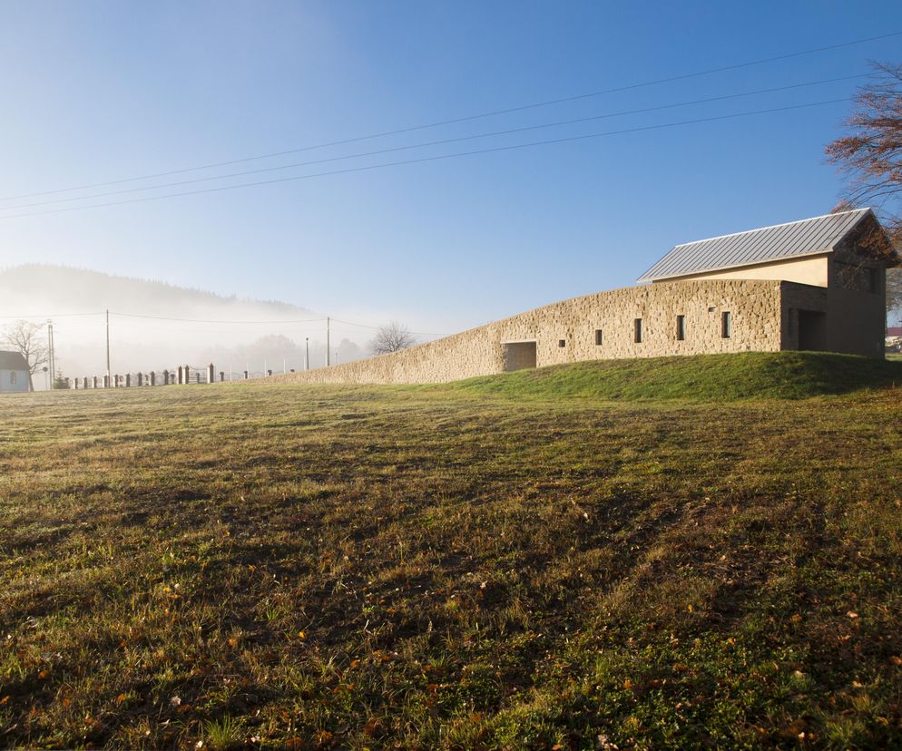 Kaplica Ostatniego Pożegnania w Rychwałdzie autorstwa Jakuba Turbasy i Bartłomieja Pyrzyka. Budynek z kamiennymi murami i spadzistym dachem, otoczony zielonym wzgórzem oraz mglistym krajobrazem, nagradzana realizacja, o której przeczytasz na Architektura Murator Plus.