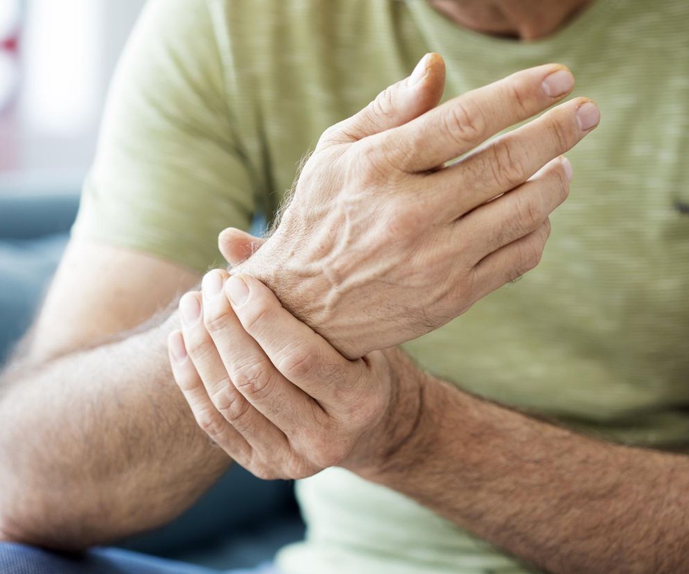 Mężczyzna w oliwkowej koszulce, trzymający się za nadgarstek, wyraźnie odczuwający ból w stawie. Widoczne dłonie, żyły i owłosienie na przedramionach. Obraz symbolizuje ukryte objawy RZS i może ilustrować artykuł na Poradnik Zdrowie o walce ciała z chorobą na lata przed pojawieniem się bólu.