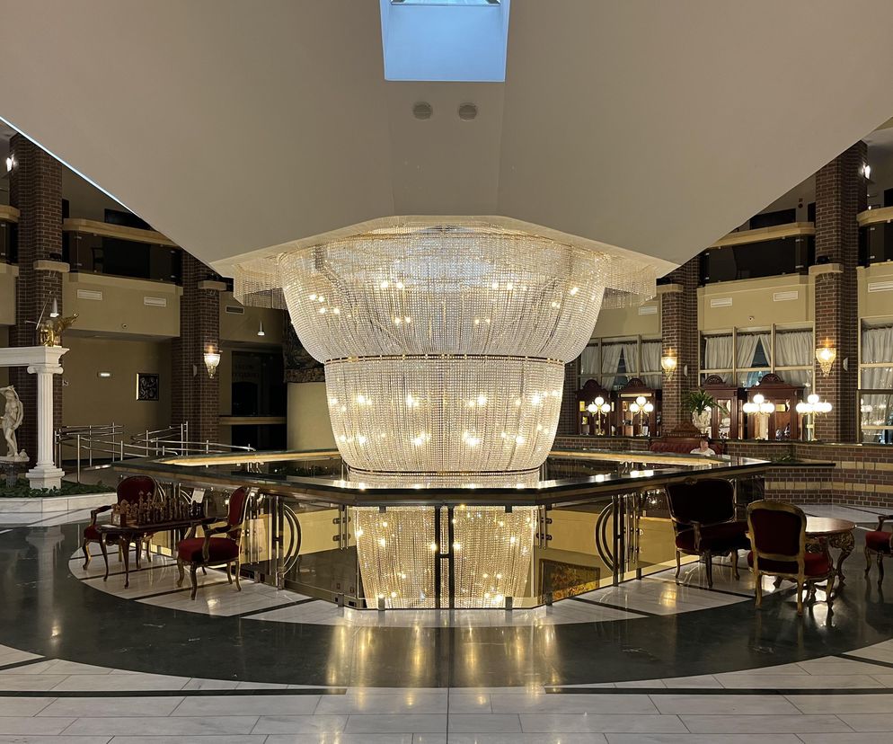 Lobby Hotelu Jana Pawła II we Wrocławiu z centralnie umieszczonym, monumentalnym żyrandolem kryształowym, spadającym przez kondygnacje i rozświetlającym wnętrze. Po lewej stronie widoczne są neoklasyczne posągi oraz kolumny, a w tle ceglane ściany. Wokół żyrandola rozmieszczone są eleganckie fotele, stoły, co podkreśla luksusowy wystrój. O postmodernistycznej architekturze czytaj na Architektura Murator Plus.