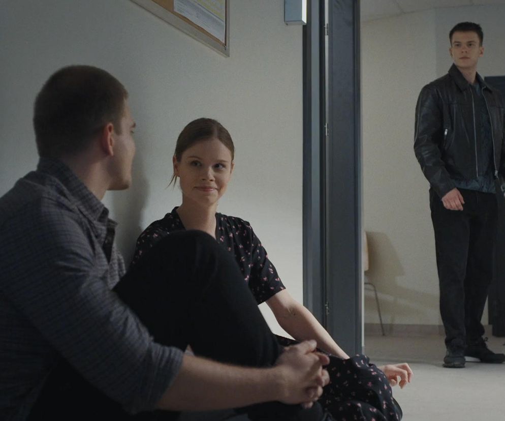 Mateusz Mostowiak i Majka siedzą na podłodze, patrząc na siebie z uśmiechem, podczas gdy Tomasz pojawia się w tle. Scena z odcinka 1882 M jak miłość, o którym więcej przeczytasz na Super Seriale.