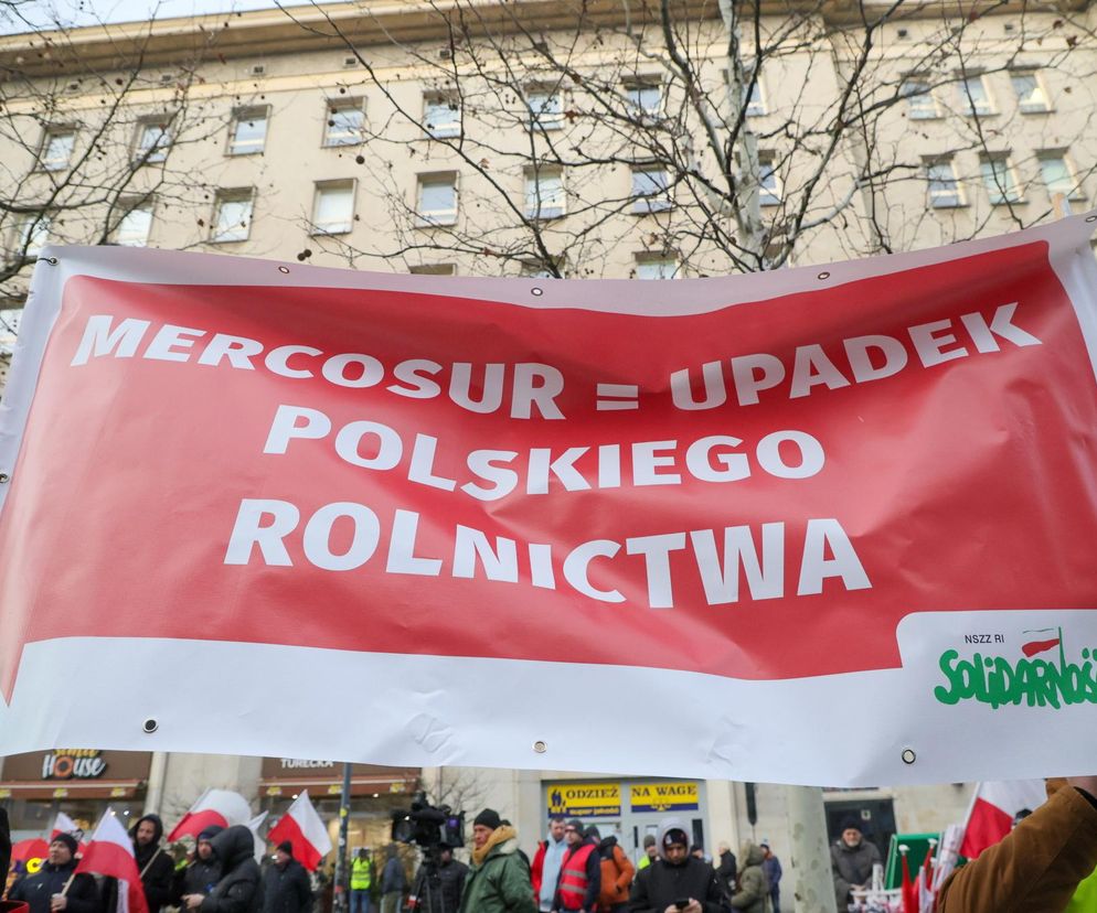 Czerwony transparent z białym napisem MERCOSUR = UPADEK POLSKIEGO ROLNICTWA i logo NSZZ RI Solidarność, niesiony podczas protestu rolników. W tle widać budynek i uczestników demonstracji z flagami Polski. Super Biznes informuje o mechanizmach ochronnych KE.
