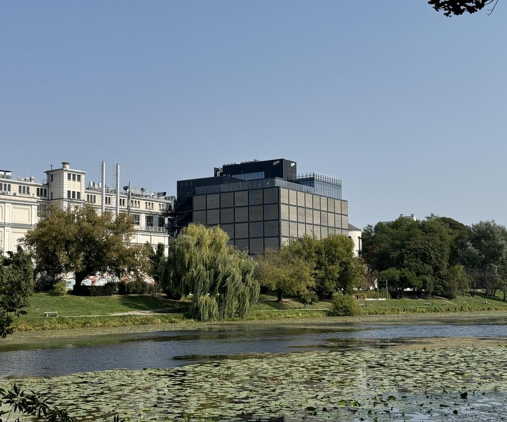 Nowoczesny, ciemnoszary budynek Muzeum Fabryki Czekolady E. Wedel, widziany od strony akwenu z liliami wodnymi. W tle, po lewej, klasyczny kremowy budynek fabryki. Drzewa na brzegu dopełniają kompozycję. Więcej o architekturze na Architektura Murator Plus.