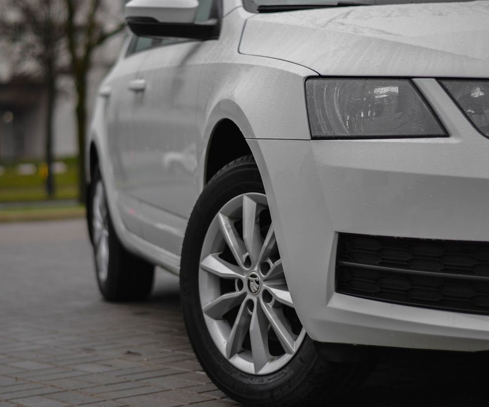 Biały samochód osobowy, prawdopodobnie Skoda Octavia, widoczny z przodu i boku, zaparkowany na ulicy w deszczowy dzień, z kroplami wody na lakierze, symbolizujący rynek aut używanych i nowych, o którym przeczytasz więcej na Super Biznes.