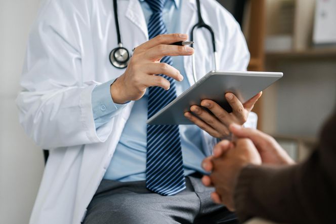 Lekarz w białym kitlu i krawacie trzymający tablet i długopis, rozmawiający z pacjentem, symbolizuje wprowadzenie e-rejestracji NFZ i cyfryzację opieki zdrowotnej od 2026 roku. Więcej o rewolucji w ochronie zdrowia przeczytasz na Poradnik Zdrowie.