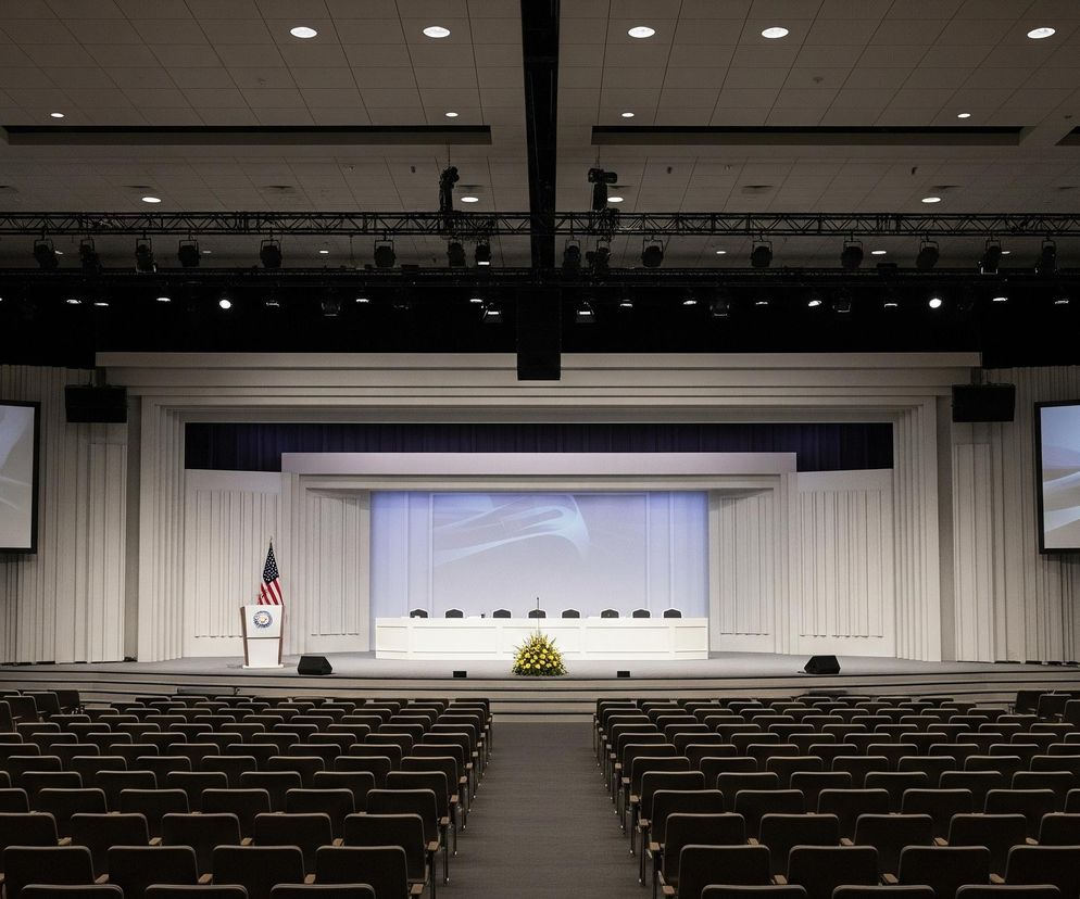 Duża sala konferencyjna, oświetlona głównie światłem z przodu sceny i projektorów, dominuje w centrum obrazu. Na środku podwyższonej sceny znajduje się biały stół prezydialny z pięcioma ciemnymi krzesłami i mikrofonami, przed którym na podłodze stoi bukiet żółtych i białych kwiatów. Po lewej stronie stołu, za białym mównicą z widoczną flagą Stanów Zjednoczonych, znajduje się amerykańska flaga, a obok mównicy, na podłodze, umieszczony jest ciemny głośnik.

Scena otoczona jest białymi, pionowymi panelami, a za stołem prezydialnym widoczny jest duży, podświetlony ekran, na którym wyświetlana jest abstrakcyjna grafika w odcieniach błękitu i szarości. Po obu stronach sceny, nad czarnymi ścianami, wiszą duże ekrany projekcyjne, wyświetlające tę samą abstrakcyjną grafikę. Sufit sali jest kasetonowy, z wbudowanymi punktowymi światłami, a poniżej niego, na czarnych kratownicach, zawieszone są reflektory i sprzęt oświetleniowy.

Sala wypełniona jest rzędami pustych, ciemnobrązowych krzeseł, ustawionych w równych rzędach na szarej wykładzinie, tworząc szerokie przejście w centrum. Krzesła mają drewniane podłokietniki i metalowe stelaże, a ich oparcia są lekko pochylone.