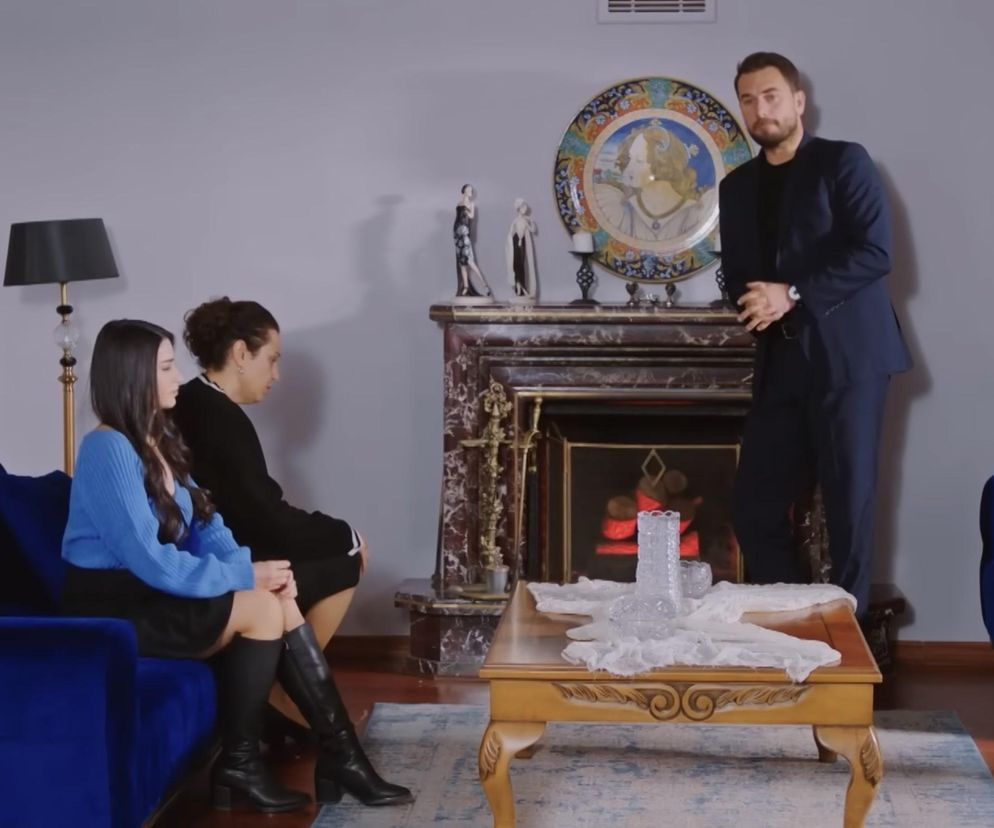 Mężczyzna w garniturze stoi przed kominkiem, patrząc na dwie kobiety siedzące na niebieskiej sofie w salonie, w kadrze z serialu „Miłość i nadzieja”. Scena sugeruje poważną rozmowę o rozwodzie Bahar i Kuzeya, którą można śledzić na portalu Super Seriale.