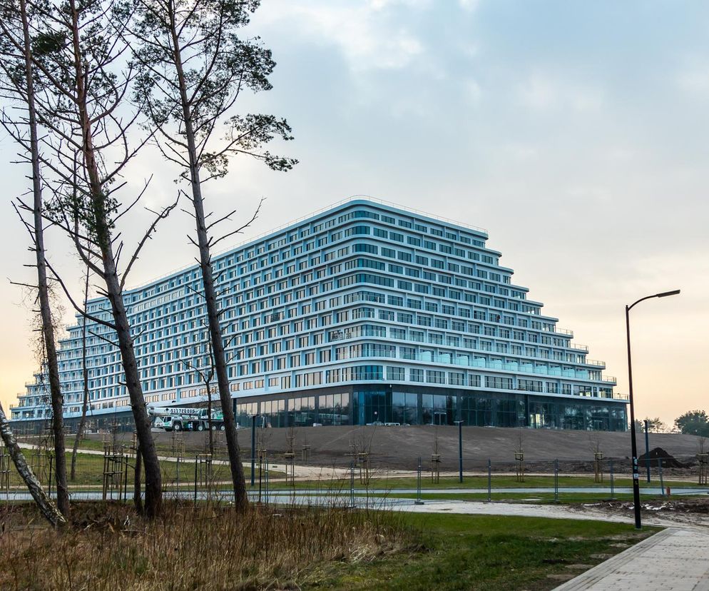 Nowoczesny Hotel Gołębiewski w Pobierowie, największy w Polsce, na tle jasnego, zachmurzonego nieba. Widoczne są liczne okna i tarasy w kaskadowej bryle budynku, otoczonego terenem budowy z młodymi drzewami i suchą trawą. O otwarciu obiektu przeczytasz na Super Biznes.
