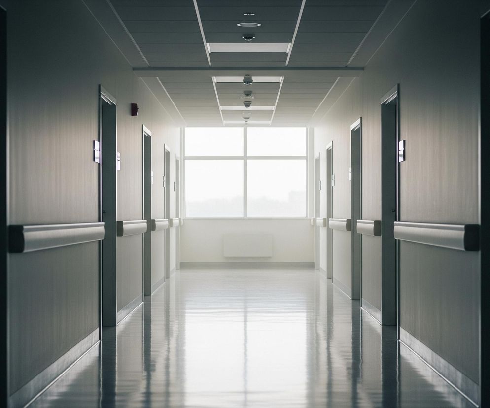 Długi korytarz z drzwiami po obu stronach prowadzi w głąb obrazu ku dużemu, prostokątnemu oknu na końcu, przez które wpada jasne światło. Ściany są jasnoszare, a na wysokości pasa biegną wzdłuż nich jasne, poziome uchwyty. Podłoga jest błyszcząca, odbijając światło i elementy otoczenia, tworząc symetryczny efekt. Sufit ma wbudowane prostokątne oprawy oświetleniowe, rozmieszczone centralnie w rzędzie, z kilkoma widocznymi okrągłymi elementami.