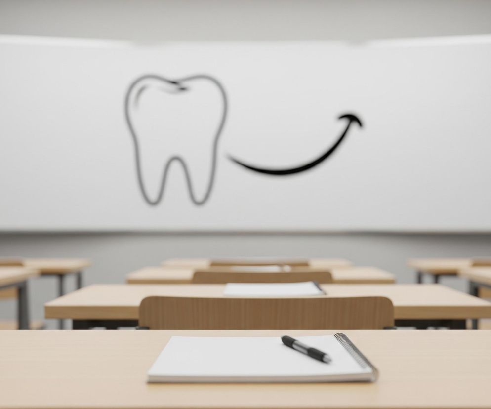 Wnętrze sali lekcyjnej z rzędami drewnianych ławek i krzeseł, ustawionych przodem do dużej, białej tablicy na ścianie. Na pierwszym planie, na najbliższej ławce, leży otwarty zeszyt z długopisem. Tablica, znajdująca się w głębi, ma narysowany kontur zęba oraz zakrzywioną strzałkę skierowaną w prawo. Ściany pomieszczenia są szare.