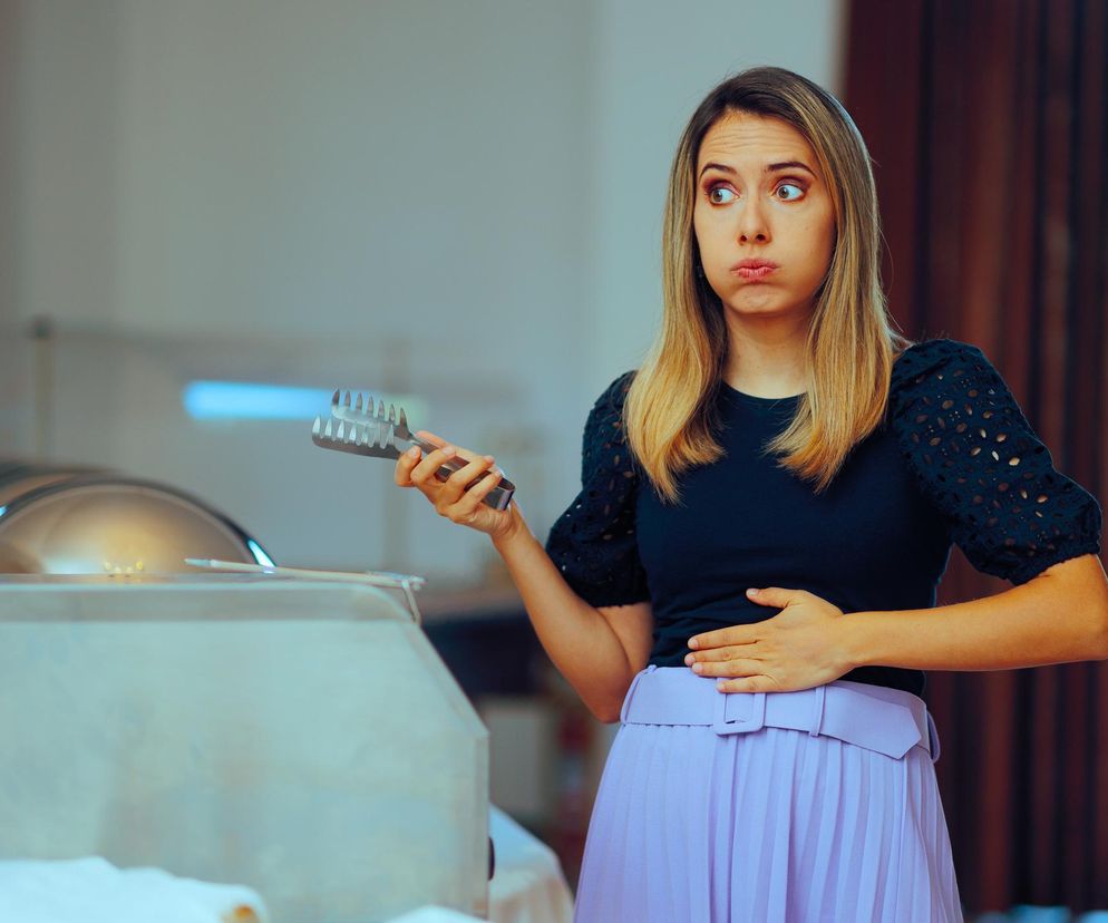 Kobieta z ręką na brzuchu, trzymająca szczypce do jedzenia, z zabawną miną, sygnalizującą burczenie w brzuchu lub dolegliwości trawienne, co podkreśla, że na Poradnik Zdrowie można znaleźć artykuły o tym, co oznaczają dźwięki z brzucha.