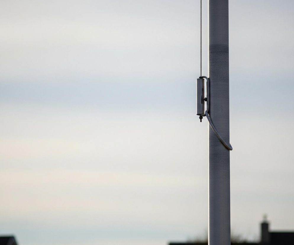 Po prawej stronie obrazu znajduje się pionowy szary maszt z metalowym mechanizmem przymocowanym do niego mniej więcej w jednej trzeciej wysokości od góry. Z mechanizmu zwisa lina, owijając się wokół masztu. Tło stanowi rozmyte niebo o jasnych, błękitno-szarych odcieniach, z niewyraźnymi ciemnymi kształtami budynków u dołu.