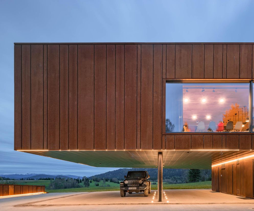 Nowoczesny Dom w Bieszczadach z elewacją z cortenu, zwisający nad podjazdem, gdzie parkuje czarny Jeep. Panoramiczne okna ukazują oświetlone wnętrze z kolorowymi krzesłami. To innowacyjna architektura wpisana w bieszczadzki krajobraz, o której przeczytasz na Architektura Murator Plus.