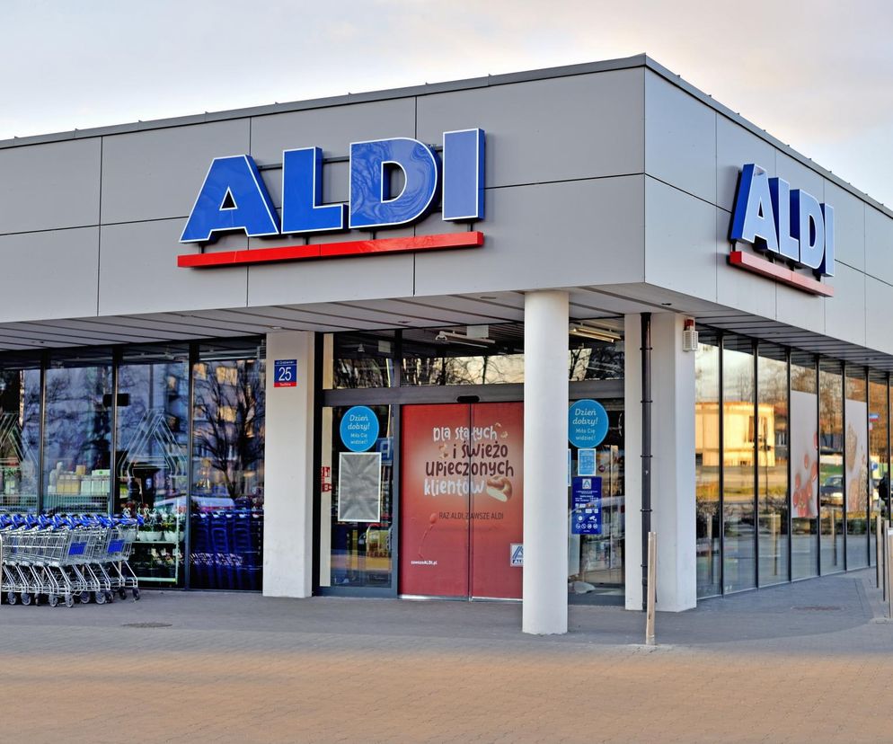 Nowoczesny budynek sklepu Aldi z charakterystycznym niebieskim logo, rzędem wózków sklepowych oraz szklaną fasadą. Wejście ozdobione jest czerwonymi naklejkami. Artykuł na Super Biznes informuje o planach ekspansji sieci dyskontów w Polsce pomimo rekordowej straty.