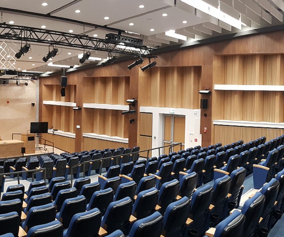 Zmodernizowana Aula Magna z nowymi, niebieskimi fotelami, drewnianymi panelami akustycznymi na ścianach i profesjonalnym oświetleniem scenicznym. Projekt remontu Auli Magna w Centrum Medycznym Kształcenia Podyplomowego w Warszawie zgłoszony do konkursu Życie w Architekturze, o którym przeczytasz na Architektura Murator Plus.