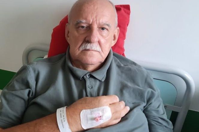 Jacek Pałasiński, były dziennikarz TVN24, w szpitalnej koszuli i z opaską identyfikacyjną na nadgarstku. Na jego dłoni widać wenflon zabezpieczony plastrem, a sam Pałasiński leży na szpitalnym łóżku, podparty czerwoną poduszką. O jego doświadczeniach ze szpitalnego jedzenia przeczytasz na Poradnik Zdrowie.