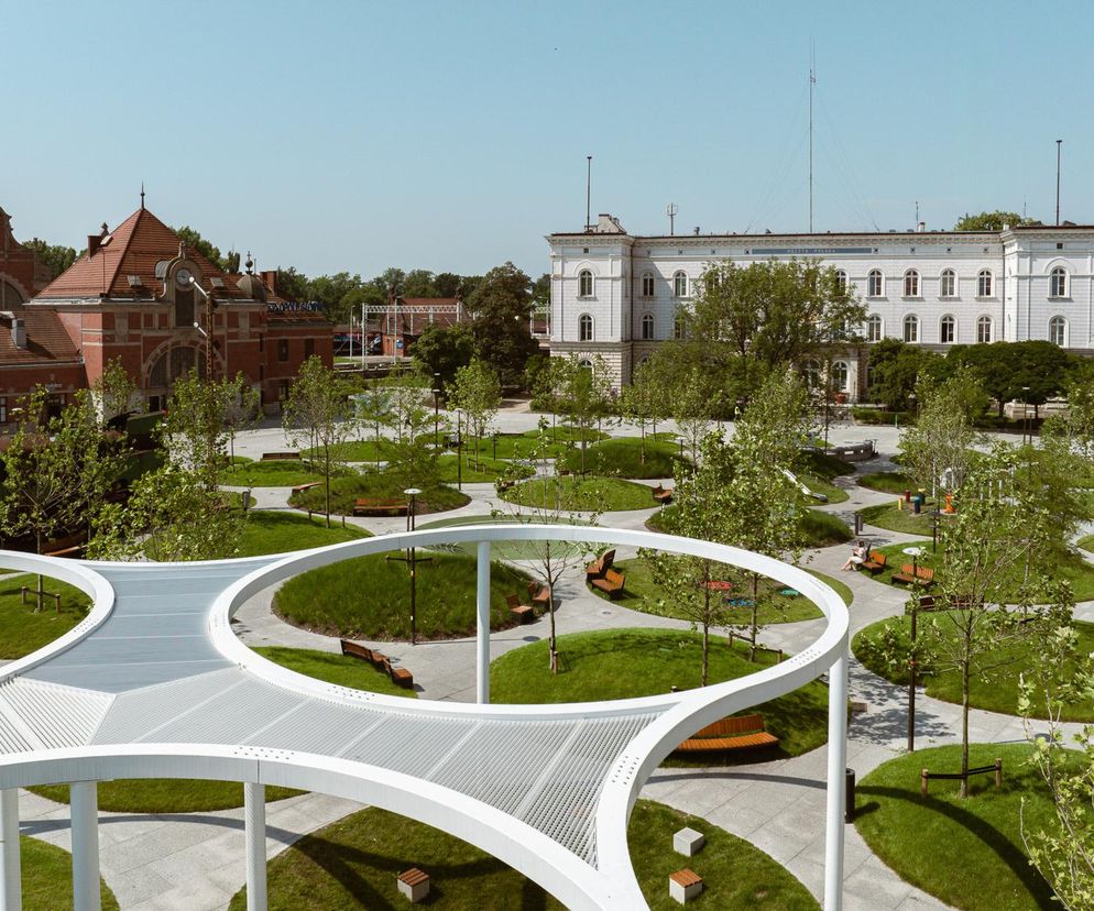 Plac im. Olgi Kory Sipowicz w Opolu, zaprojektowany przez SN Architekci, z charakterystycznymi kolistymi rabatami traw i ścieżkami. Na pierwszym planie biała, nowoczesna pergola, w tle Dworzec Główny i zabytkowy budynek poczty. Architektura Murator Plus prezentuje zgłoszenie do konkursu Życie w Architekturze 2025.