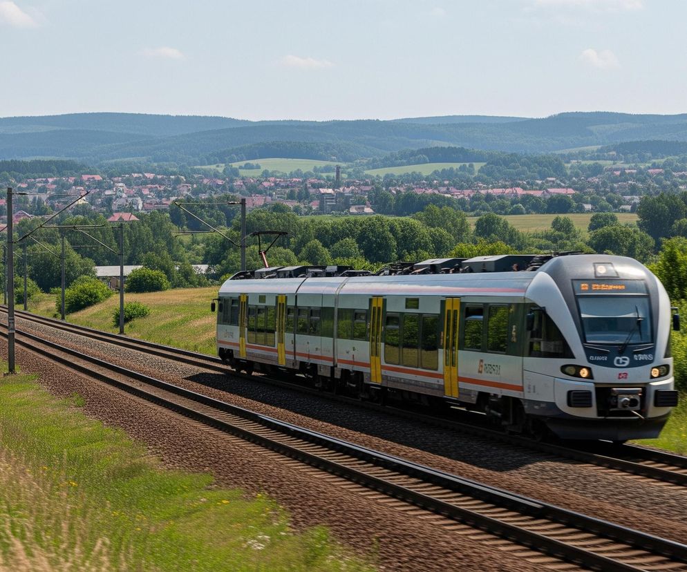 Nowoczesny pociąg osobowy w kolorze biało-szarym z żółtymi drzwiami i pomarańczowymi pasami po bokach porusza się po torach, widoczny od prawej strony obrazu w kierunku centrum. Torowisko składa się z trzech równoległych szyn na podsypce żwirowej, biegnących w lewo i w głąb obrazu, obok zielonego trawnika porośniętego bujną trawą i pojedynczymi krzewami. W tle rozciąga się pagórkowaty krajobraz z rozległymi terenami zielonymi, pokrytymi lasami i małą miejscowością z dachami budynków w odcieniach czerwieni i brązu. Niebo jest błękitne z niewielką ilością białych chmur.