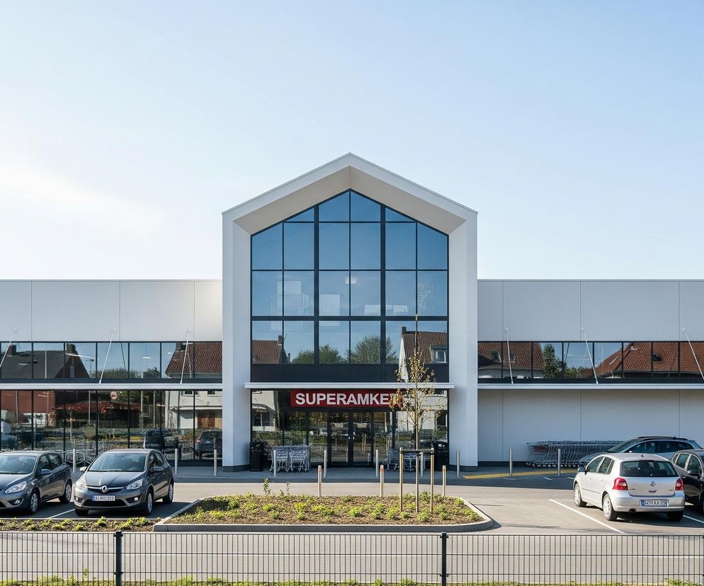 Centralną część obrazu zajmuje nowoczesny budynek w kolorze białym, z dużymi oknami odbijającymi otoczenie i niebo. Fasada wejściowa ma kształt trójkątnego szczytu z całkowicie przeszkloną powierzchnią, pod którą widnieje czerwony neon z napisem SUPERAMKET. Przed budynkiem rozciąga się przestronny parking, na którym zaparkowanych jest kilka samochodów różnych marek i kolorów, a także ustawione są rzędy wózków sklepowych. Na pierwszym planie widoczny jest czarny metalowy płot oddzielający teren parkingu od niewidocznego obszaru poza kadrem, a także zielone klomby z młodymi drzewkami i krzewami. W tle, po bokach budynku, zarysowują się dachy i fasady okolicznych domów, niektóre z cegły i czerwonymi dachówkami, pod jasnym, bezchmurnym niebem.