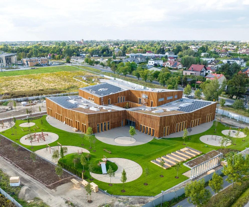 Energoefektywne przedszkole w Regułach z drewnianymi elewacjami i zielonym dachem, widok z lotu ptaka. Na dachu panele słoneczne, a wokół budynku nowoczesny plac zabaw z piaskownicami i trawą. Więcej o architekturze tego typu przeczytasz na Architektura Murator Plus.