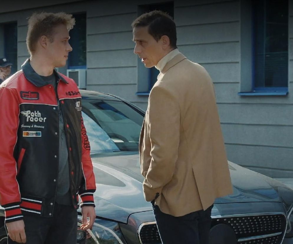 Dominik (Bartosz Surówka) w czerwono-czarnej kurtce baseballowej rozmawia z ojcem Walatem (Szymon Mysłakowski) w beżowej marynarce i golfie, stojąc przy ciemnym samochodzie przed budynkiem. Za nimi w tle widać policjanta. Obaj mężczyźni, postaci z serialu M jak miłość, omawiają strategię przesłuchania, co świadczy o skomplikowanej sytuacji Dominika, o czym można przeczytać na Super Seriale.