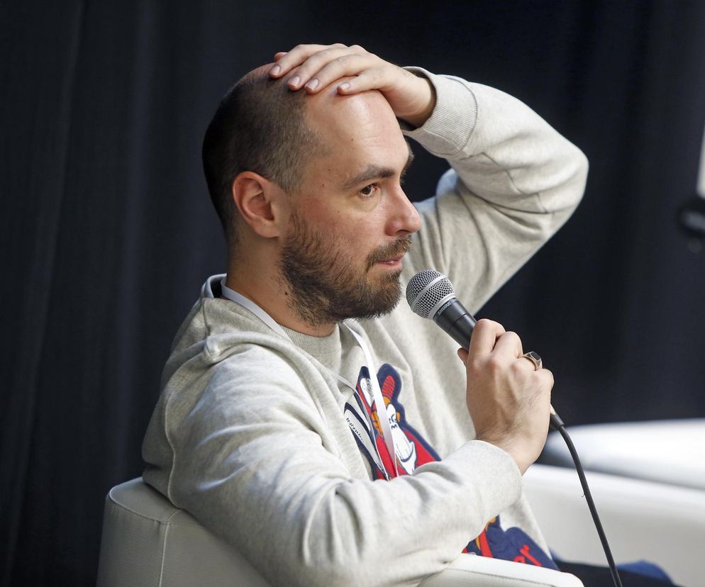 Pisarz Jakub Żulczyk z mikrofonem podczas publicznego wystąpienia. Mężczyzna w jasnej bluzie z kapturem, z brodą, łapie się za głowę, zamyślony lub zmartwiony. Osobista relacja Żulczyka o psie, który zmarł w pociągu, dostępna jest na naszym portalu.