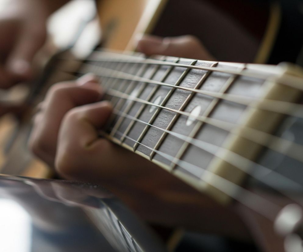 Główne ujęcie przedstawia fragment gry na gitarze akustycznej. Centralnie widoczny jest gryf gitary z metalowymi progami i biało-brązowymi strunami, rozciągającymi się ukośnie przez kadr. Na gryfie spoczywa dłoń z palcami układającymi się do akordu, zaś w tle, nieco rozmazana, widać drugą dłoń uderzającą w struny. Kolory są ciepłe, dominują brązy drewna gitary, biele strun oraz jasne odcienie skóry dłoni, z jasnym, rozmytym tłem.