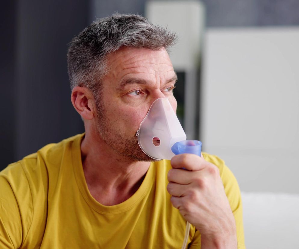 Mężczyzna w średnim wieku z siwymi włosami, w żółtej koszulce, używa inhalatora, trzymając maskę na twarzy. Jego wyraz twarzy sugeruje skupienie lub zaniepokojenie. Zdjęcie ilustruje problem duszności przy POChP, o której przeczytasz więcej na portalu Poradnik Zdrowie.