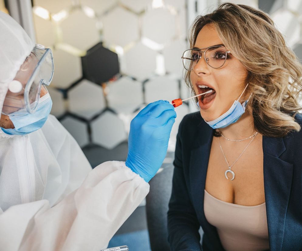 Osoba w ochronnym kombinezonie, okularach i masce pobierająca wymaz z gardła kobiety z odsłoniętymi ustami, w okularach i rozpuszczonych włosach. Na Poradnik Zdrowie znajdziesz informacje o testach na koronawirusa.