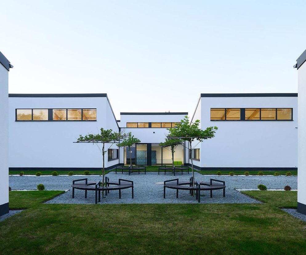 Nowoczesny kampus modułowej szkoły podstawowej nr 17 w Rudzie Śląskiej, z białymi elewacjami, czarnymi detalami i zielonymi dziedzińcami. Widoczne krzewy, ławki i drzewka. Więcej o architekturze placówek edukacyjnych przeczytasz na Architektura Murator Plus.