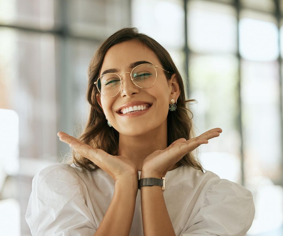 Młoda kobieta z długimi, brązowymi włosami i w okrągłych okularach radośnie się śmieje, zamykając oczy i opierając dłonie pod brodą, co symbolizuje relaks i dobry nastrój. Na portalu Poradnik Zdrowie znajdziesz więcej informacji o redukcji stresu terapią śmiechem.