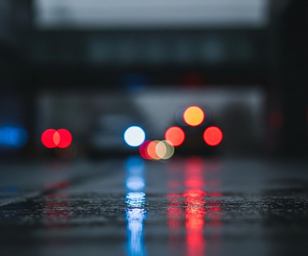 Mokry asfalt w pierwszym planie odbija różnokolorowe światła, tworząc wydłużone smugi czerwieni i błękitu. Na drugim planie widoczne są rozmyte, okrągłe źródła światła – głównie czerwone i pomarańczowe, z pojedynczymi białymi i niebieskimi akcentami – sugerujące pojazdy. Tło jest ciemne i zamazane, co wskazuje na nocne ujęcie miejskiej przestrzeni, być może tunelu lub wiaduktu.