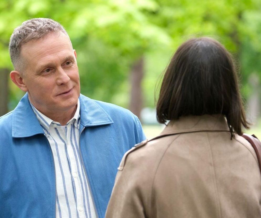 Artur Rogowski (Robert Moskwa) rozmawia z Agnes (Amanda Mincewicz) w parku, na tle zielonych drzew, spoglądając na nią z delikatnym uśmiechem. Mężczyzna jest w niebieskiej kurtce i pasiastym T-shircie, kobieta odwrócona tyłem, ma ciemne włosy i beżowy płaszcz. Więcej o ich relacji przeczytasz na Super Seriale.