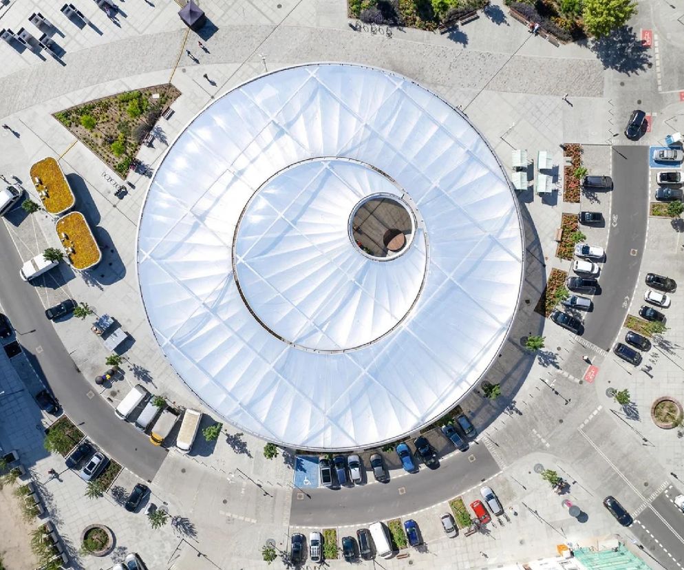 Widok z lotu ptaka na Rynek Łazarski w Poznaniu, z centralnie umieszczonym, okrągłym zadaszeniem hali targowej, otoczonym przestrzenią miejską z parkingami i zielenią, idealnie wpisującym się w kontekst artykułu o Życiu w Architekturze 2025. Więcej o projekcie na Architektura Murator Plus.