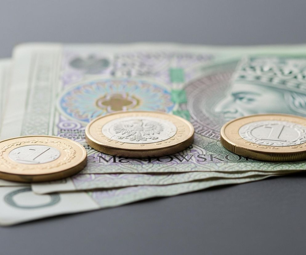 Trzy bimetaliczne monety o złotych brzegach i srebrnym środku ułożone są na tle kilku banknotów. Monety leżą w rzędzie, ukośnie do krawędzi banknotów, z wyraźnym numerem 1 na środkowej części dwóch z nich oraz stylizowanym orłem na trzeciej monecie. Banknoty, częściowo zielone i fioletowe, przedstawiają wizerunek postaci w koronie oraz fragmenty numerów seryjnych. Wszystko to spoczywa na ciemnoszarym, gładkim tle, które w górnej części zdjęcia jest lekko rozmyte.