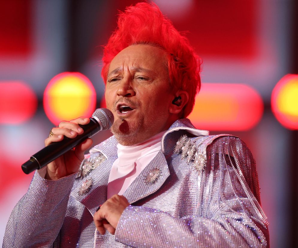 Michał Wiśniewski w trakcie występu, z charakterystycznymi czerwonymi włosami, śpiewający do mikrofonu. Ma na sobie błyszczącą marynarkę, a w tle widać rozmazane czerwone i pomarańczowe światła. O walce z alkoholizmem można przeczytać na Poradnik Zdrowie.