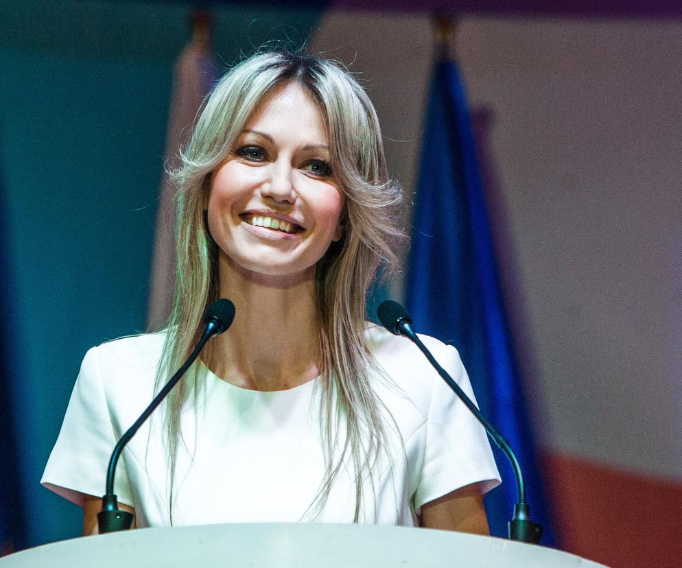 Magdalena Ogórek, prezenterka, uśmiecha się podczas wystąpienia, stojąc za mównicą z mikrofonami. Ma na sobie białą bluzkę, a jej jasne, długie włosy układają się swobodnie. W tle widać zarysy flag w odcieniach niebieskiego. Więcej o życiu osobistym Magdaleny Ogórek przeczytasz na Poradnik Zdrowie.