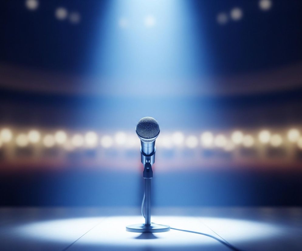 Centralnie na obrazie znajduje się srebrny mikrofon na statywie, oświetlony jasnym, niebieskim snopem światła padającym z góry. Mikrofon stoi na ciemnej, drewnianej scenie, która odbija subtelne światło. Tło stanowi rozmazana przestrzeń z rzędami jasnych, rozmytych świateł, tworzących ciepłe, bursztynowe refleksy, sugerujące widownię w ciemnym pomieszczeniu.