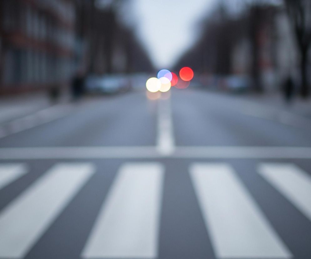 Na pierwszym planie widoczna jest rozmyta zebra, czyli pasy dla pieszych, w odcieniach bieli i szarości, przecinająca drogę. Droga, również rozmyta, prowadzi w głąb kadru, z białą, przerywaną linią pośrodku. W tle, w oddali, znajdują się rozmyte światła samochodowe: czerwone, niebieskie i żółte. Po obu stronach drogi widać rozmyte sylwetki budynków i drzew, a także pojedyncze, nieostre znaki drogowe.
